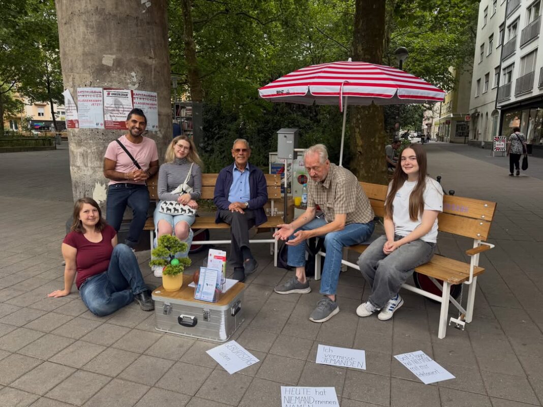 Menschen sitzen unter einem Sonnenschirm auf der Plauderbank am Kopernikusplatz